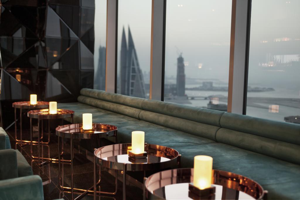 The Domain Bahrain Hotel And Spa - Adults Friendly 16 Years Plus Манама Экстерьер фото The photo depicts a stylish lounge area with a modern aesthetic. There are several circular tables made of metallic materials, each topped with a small glowing candle. A long, plush seating area in a teal color runs along the window, offering a view 